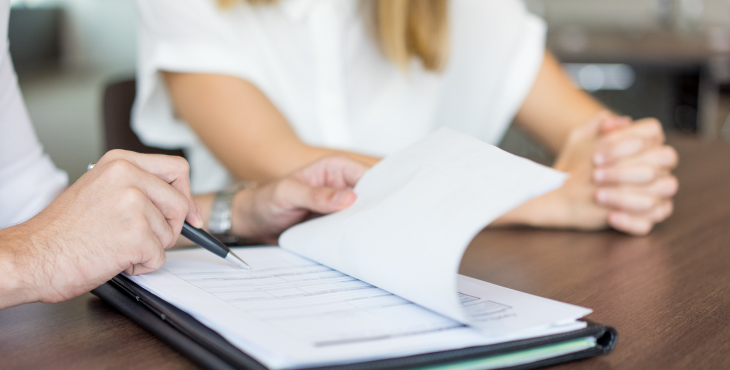 Dos personas sentadas en una mesa. Una de las personas está sosteniendo un bolígrafo y parece estar señalando o escribiendo en un documento que está sobre la mesa. La otra persona tiene las manos entrelazadas y está observando el documento. El documento parece ser un formulario o un contrato, ya que tiene varias líneas y cuadros para llenar. La imagen sugiere una situación de revisión o firma de documentos, lo cual es común en contextos laborales, legales o administrativos.