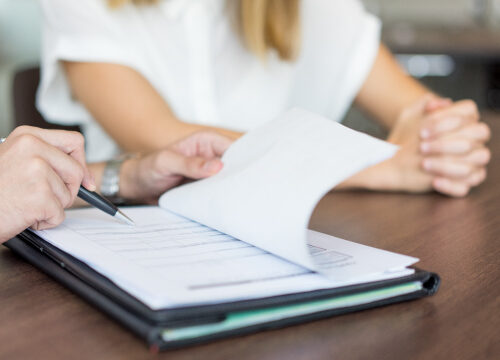 Dos personas sentadas en una mesa. Una de las personas está sosteniendo un bolígrafo y parece estar señalando o escribiendo en un documento que está sobre la mesa. La otra persona tiene las manos entrelazadas y está observando el documento. El documento parece ser un formulario o un contrato, ya que tiene varias líneas y cuadros para llenar. La imagen sugiere una situación de revisión o firma de documentos, lo cual es común en contextos laborales, legales o administrativos.