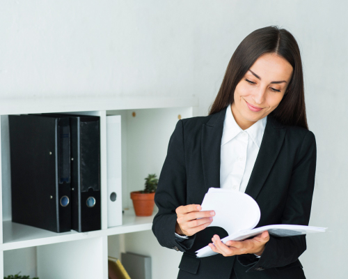 Una persona vestida con un traje formal negro y camisa blanca está revisando documentos en una oficina referente al cambio de dirección. En el fondo, hay un estante blanco con archivadores y una planta en maceta.