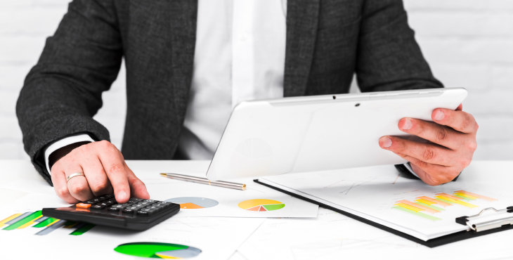 Un hombre en traje está usando una calculadora y una tableta en una mesa llena de documentos y gráficos, calculando la declaración de impuestos (Taxes).