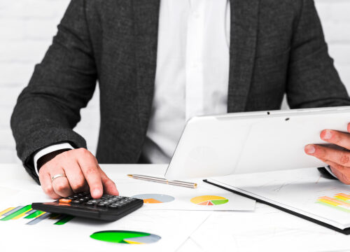 Un hombre en traje está usando una calculadora y una tableta en una mesa llena de documentos y gráficos, calculando la declaración de impuestos (Taxes).