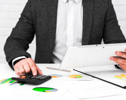 Un hombre en traje está usando una calculadora y una tableta en una mesa llena de documentos y gráficos, calculando la declaración de impuestos (Taxes).