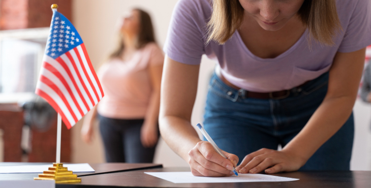 Una mujer está concentrada llenando un formulario sobre una mesa. En la mesa hay una pequeña bandera de Estados Unidos, sugiriendo un trámite migratorio. En el fondo, se ve otra persona de pie, lo que añade una dinámica de interacción y apoyo en el proceso. La imagen evoca una escena de diligencia y profesionalismo, enmarcada en un contexto cívico o administrativo.