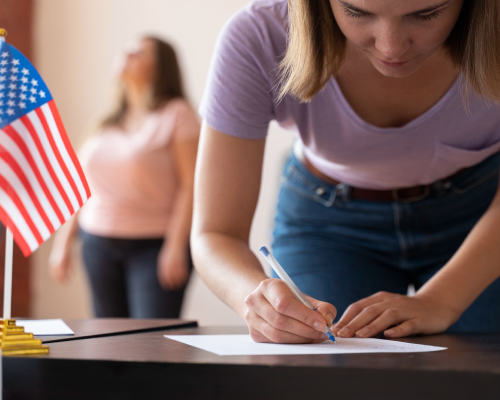 Una mujer está concentrada llenando un formulario sobre una mesa. En la mesa hay una pequeña bandera de Estados Unidos, sugiriendo un trámite migratorio. En el fondo, se ve otra persona de pie, lo que añade una dinámica de interacción y apoyo en el proceso. La imagen evoca una escena de diligencia y profesionalismo, enmarcada en un contexto cívico o administrativo.